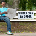 A non-white man sits on a bench in a park reserved for whites