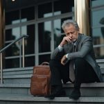 A middle-aged man in a business suit sits on the steps of his office in a depressed state. Ageism concept