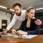 A bearded male Manager harasses his colleague a girl who works at a laptop in the office